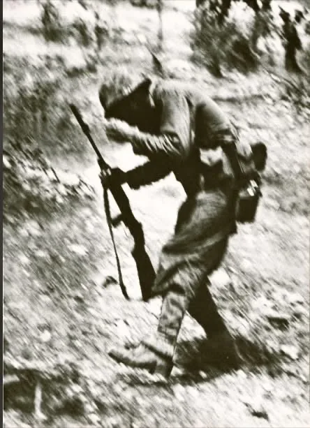 A grainy black-and-white photo shows a soldier in uniform, crouched and moving through rough terrain while carrying a rifle…