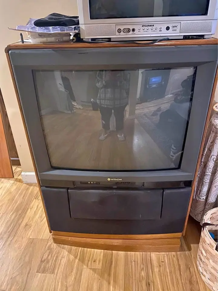 A person stands in front of a large, old Hitachi CRT television, taking a photo of their reflection in the screen. The TV is…