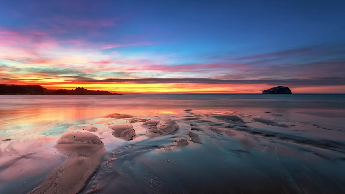 This stunning shot captures Tantallon Castle's ruin, Bass Rock in the distance, and a serene sunset over a beach. During…