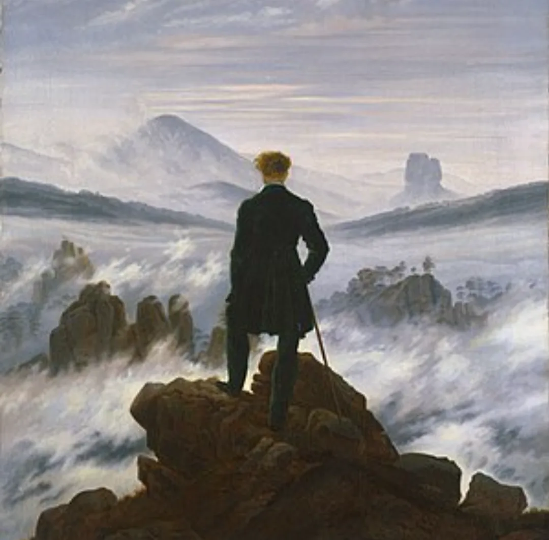 A man in a dark coat stands atop a rocky peak overlooking a vast, misty mountain landscape with distant peaks visible…