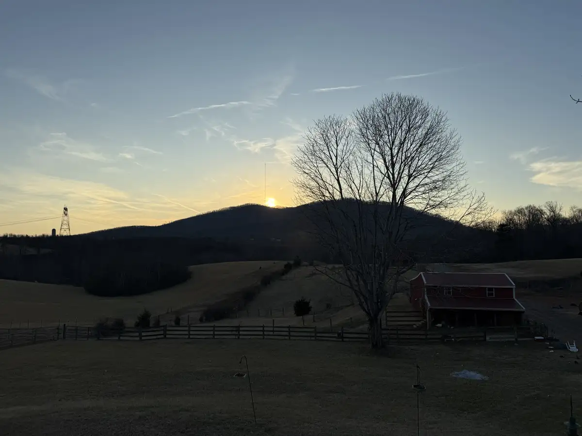 A serene rural sunset photo shows a red barn, bare trees, and rolling hills with the sun setting behind a distant mountain…