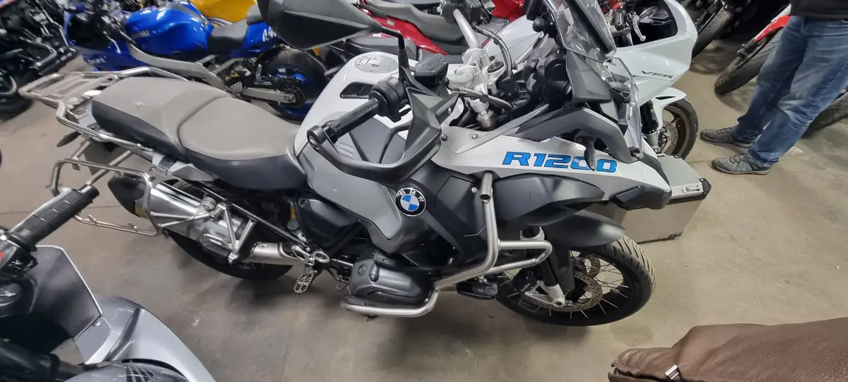 A silver BMW R1200 adventure motorcycle is parked indoors among other bikes, with its model name 'R1200' visible on the…