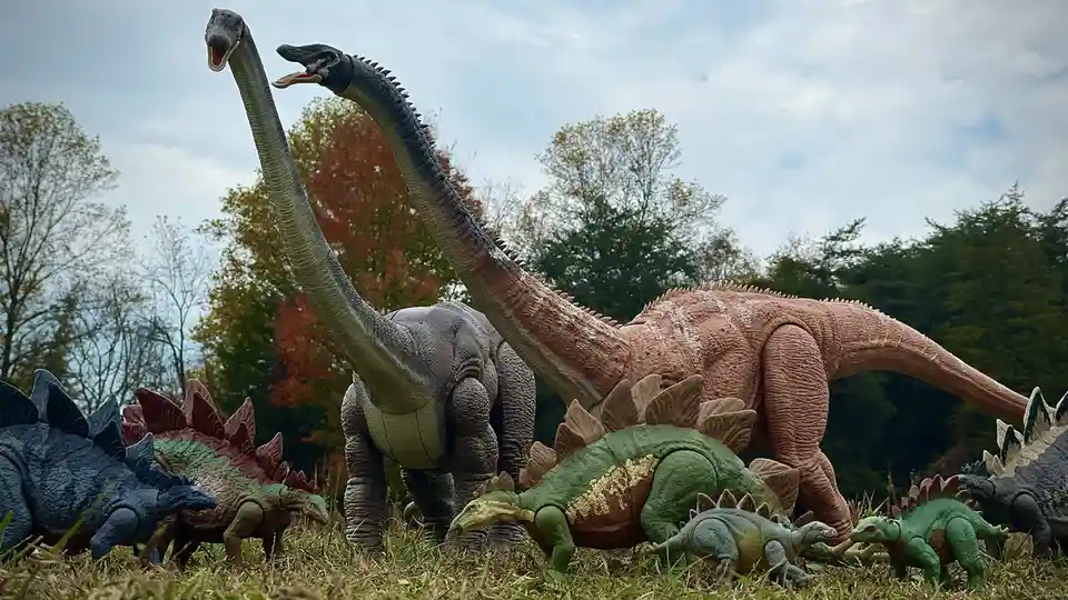 A photo of various dinosaur toys arranged outdoors on grass, featuring two large long-necked dinosaurs (likely brachiosaurs)…