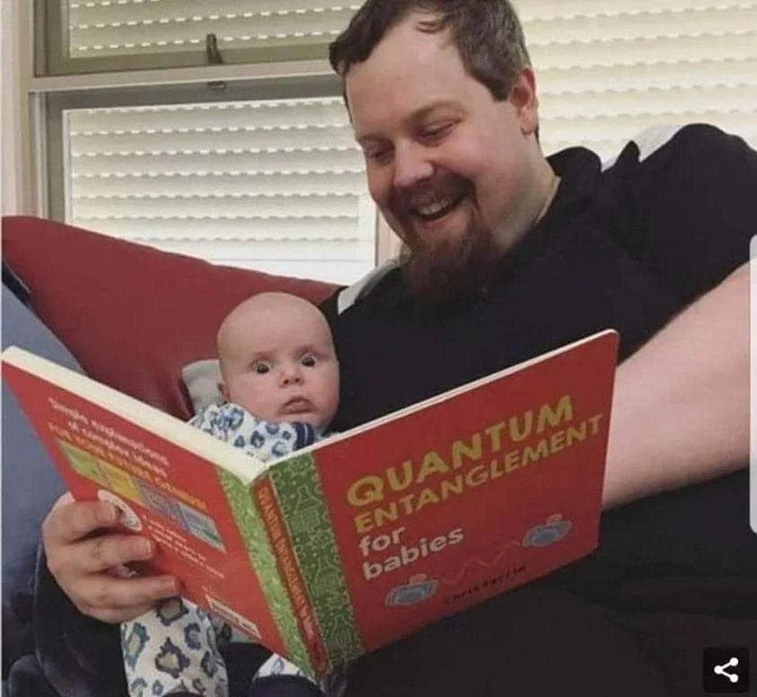 A smiling adult with a beard sits on a red couch holding a baby, who stares blankly ahead. The adult holds an open red book…