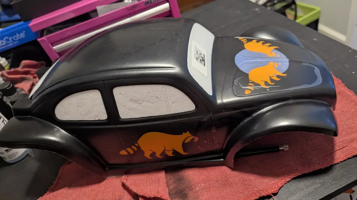 A black toy car with orange raccoon decals on its side and hood sits on a red cloth. The car has a white sticker on its rear…