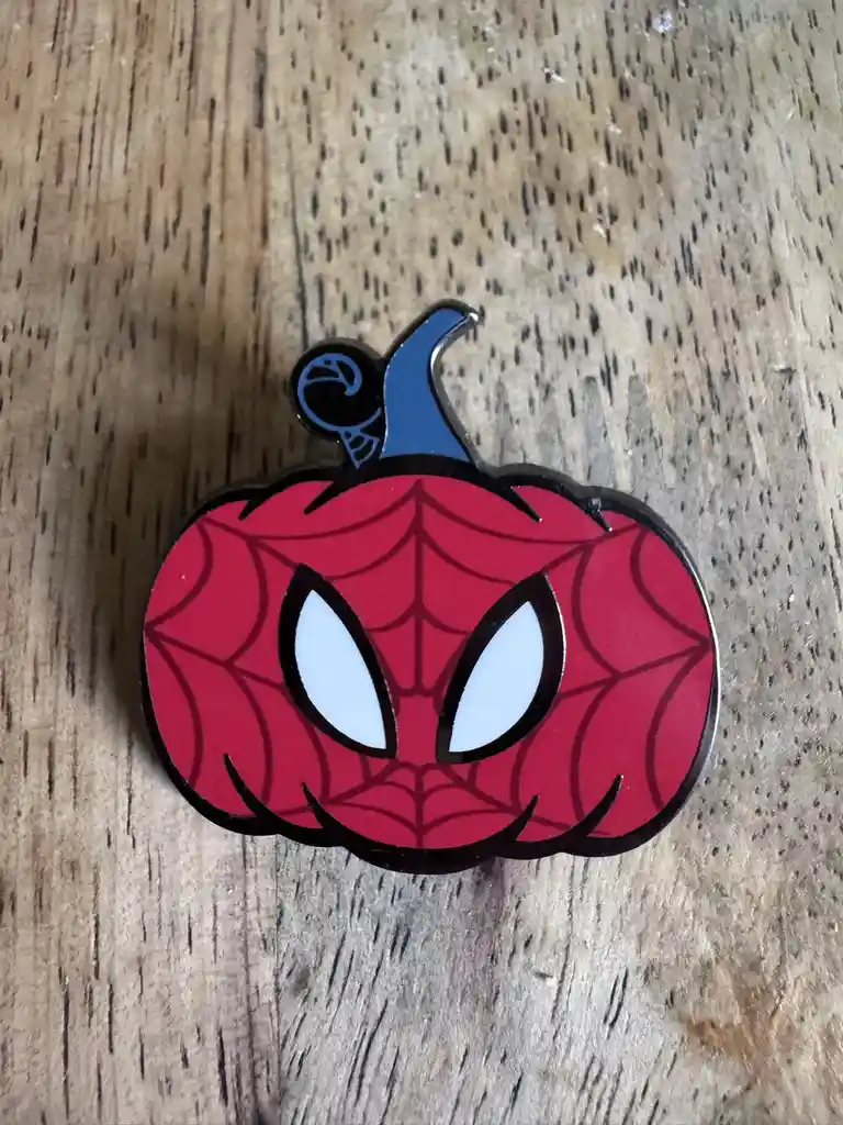 A red enamel pin shaped like a jack-o'-lantern with Spider-Man's iconic web pattern and large white eyes, topped with a blue…