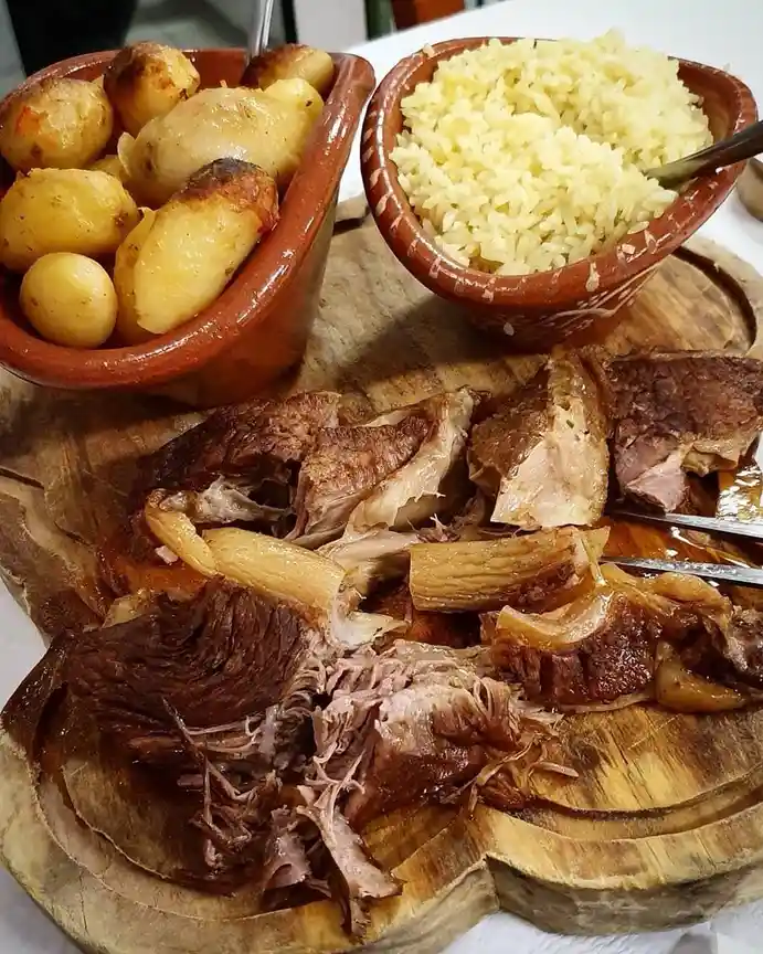 A close-up photo of a rustic meal featuring shredded roasted meat on a wooden platter, accompanied by two clay bowls—one…
