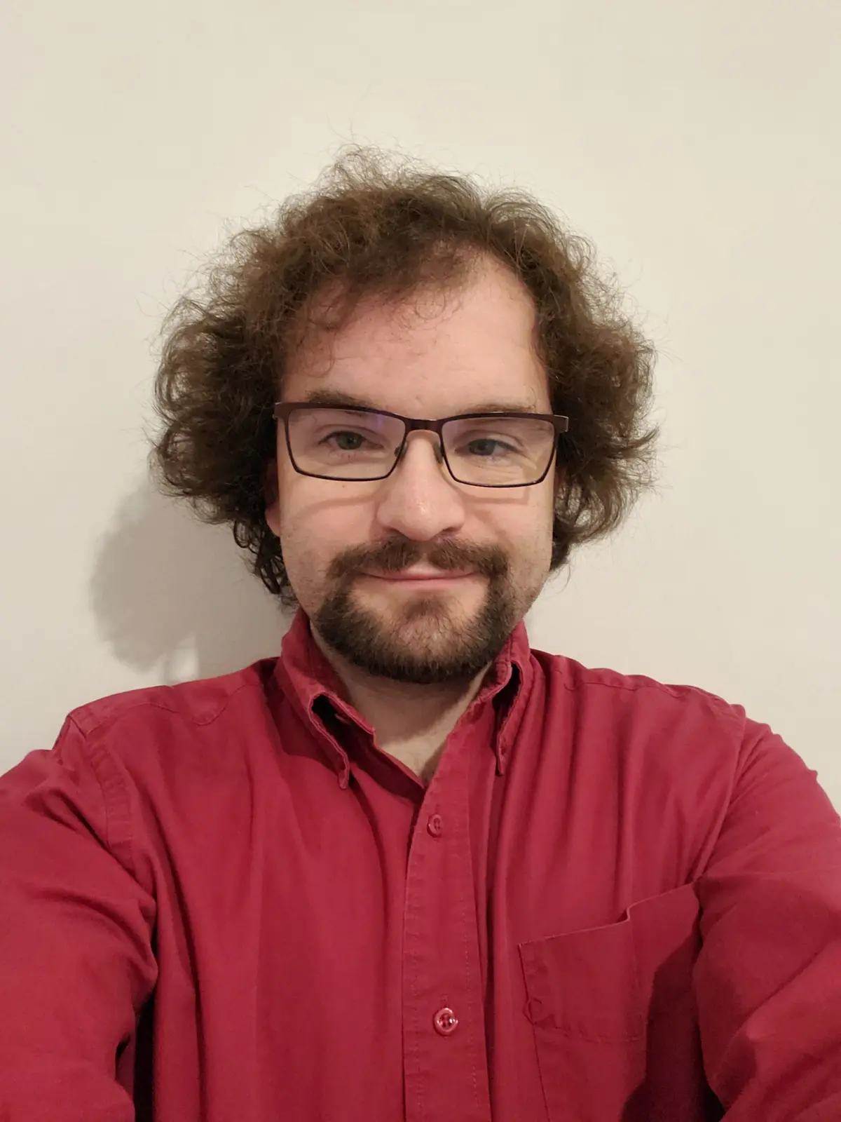 A man with curly brown hair, glasses, and a beard wears a red button-up shirt and smiles slightly while looking at the…