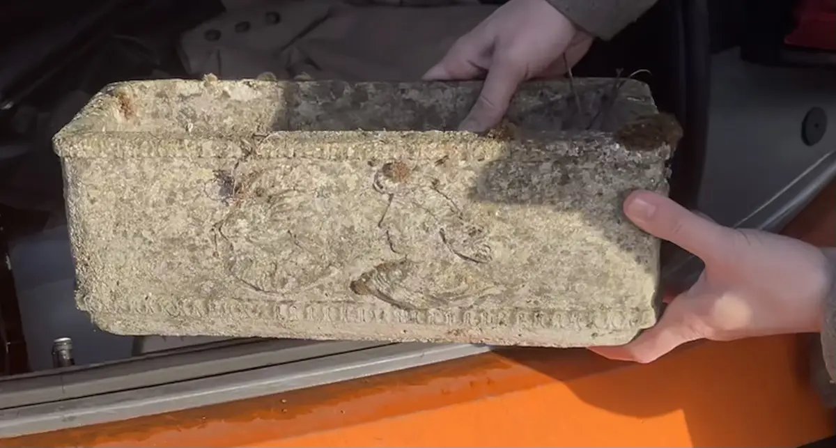 Two hands hold up a weathered, rectangular stone coffin with faint carvings and inscriptions, partially covered in dirt and…