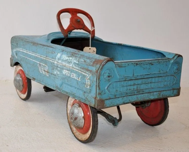 A vintage, weathered blue metal pedal car with red wheels and steering wheel, showing significant rust and chipped paint…
