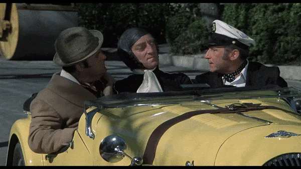Two men in a yellow convertible car, one pointing ahead as they drive past a large house with 'BURT REYNOLDS' signs above…