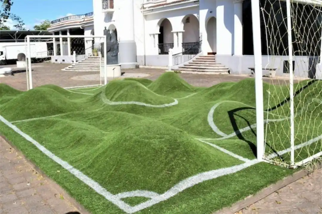 an outdoor area with artificial green turf marked with white lines to resemble a soccer field, but the turf is unevenly…