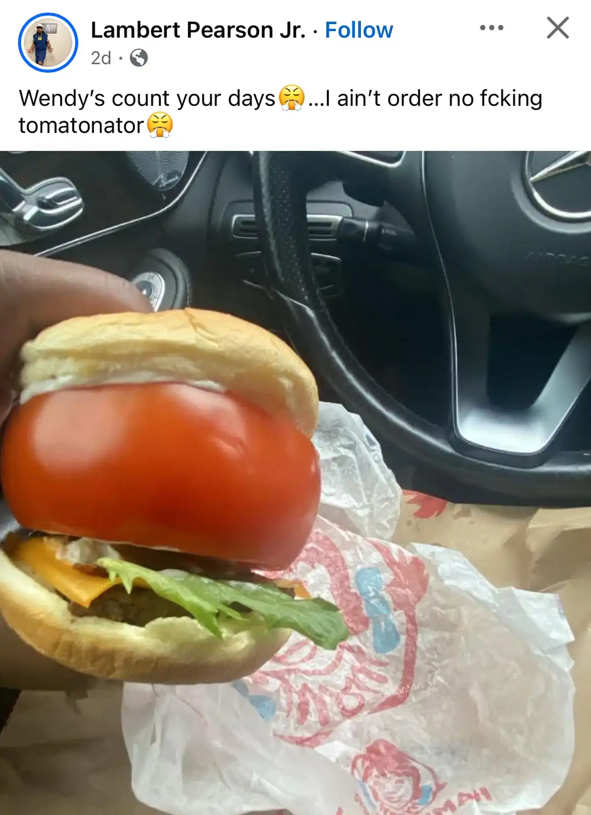 A hand holds a Wendy's hamburger with a large, whole tomato as the top bun, sitting inside a car. The burger wrapper is…
