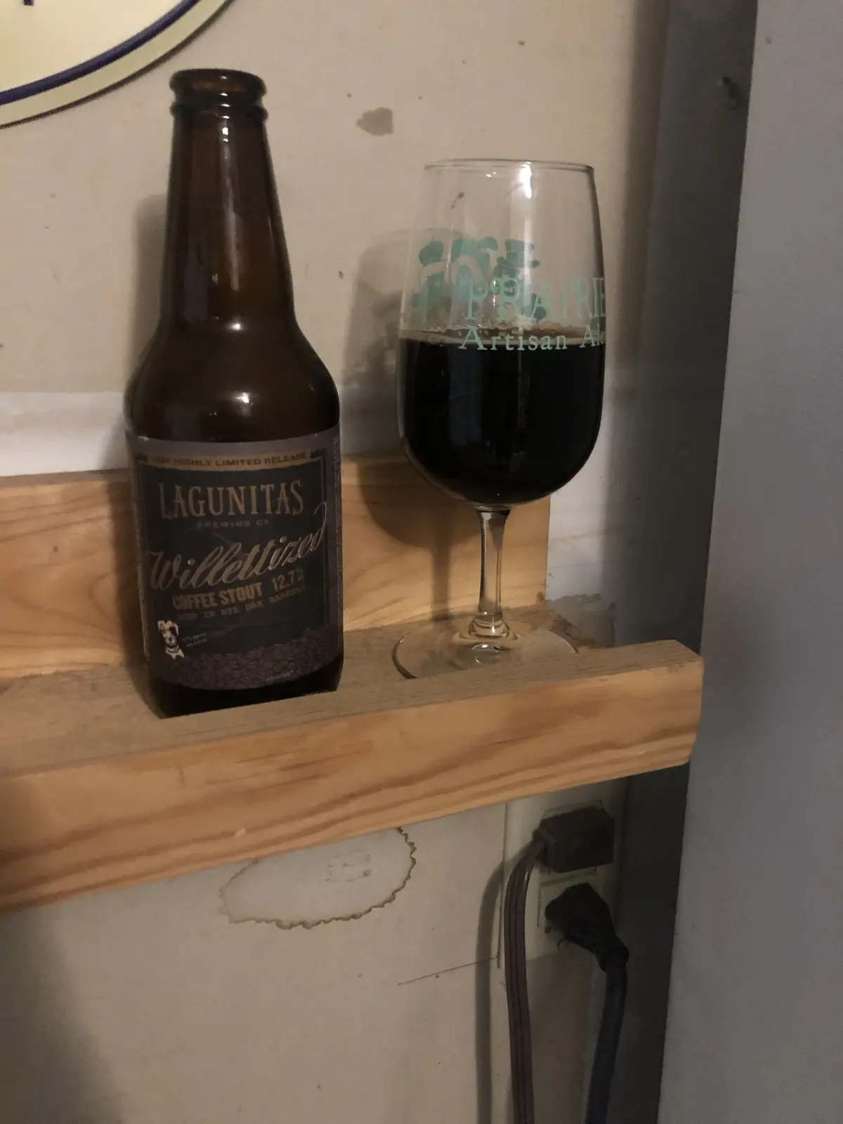 A brown bottle of Lagunitas Willettized Coffee Stout sits next to a glass of dark beer on a wooden shelf, with the word…