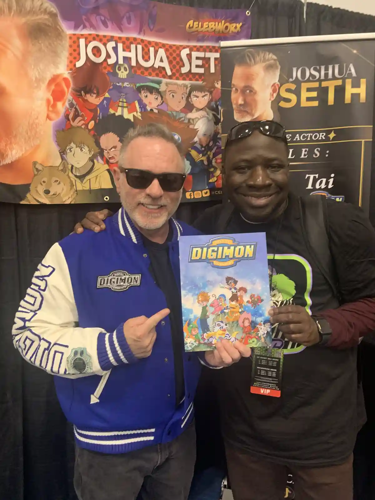 Two men pose for a photo at a convention, one wearing a blue and white Digimon varsity jacket and pointing at a Digimon…
