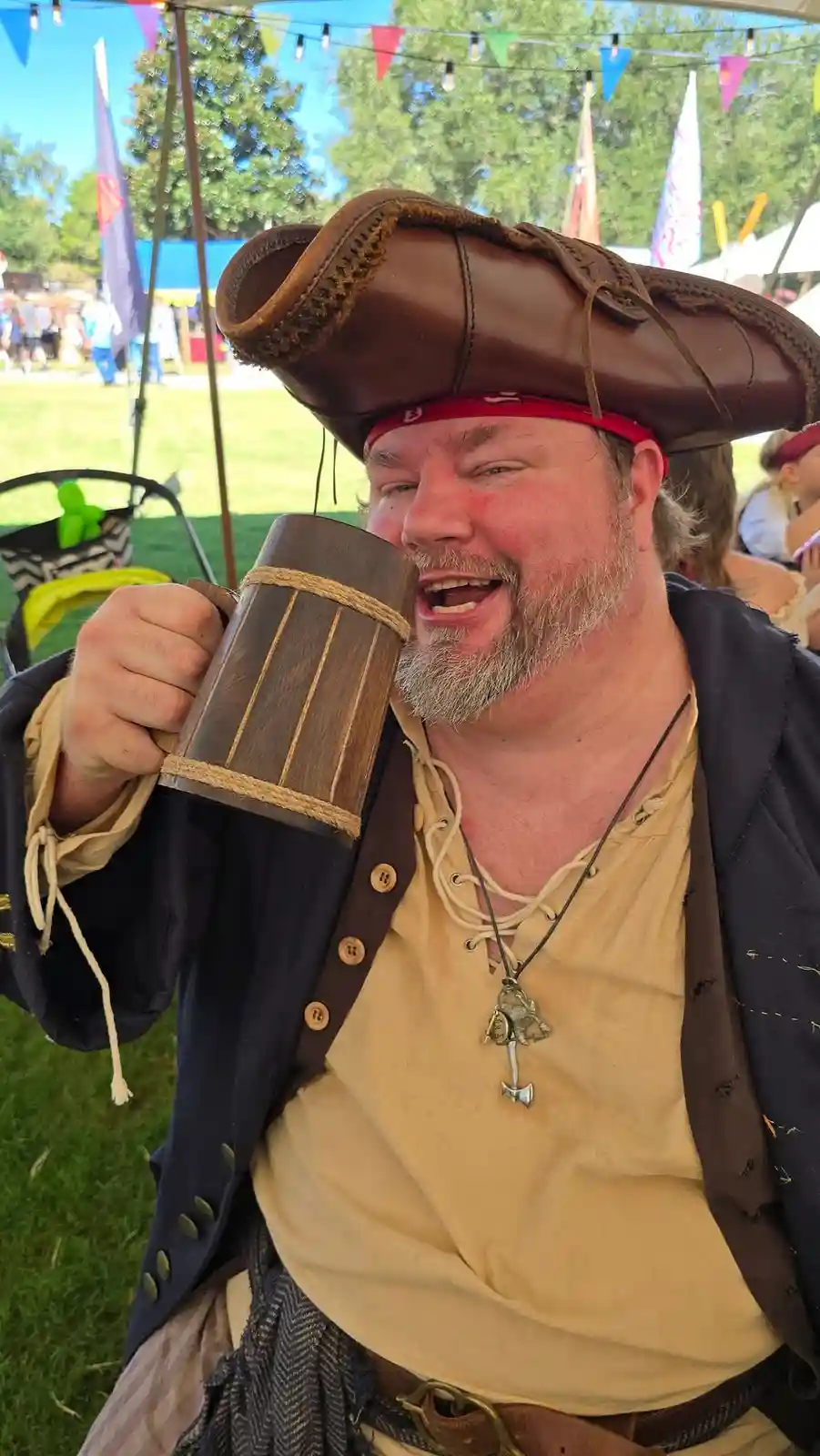 A man dressed as a pirate in a brown tricorn hat and red bandana, holding a wooden mug, smiles broadly at the camera. He…