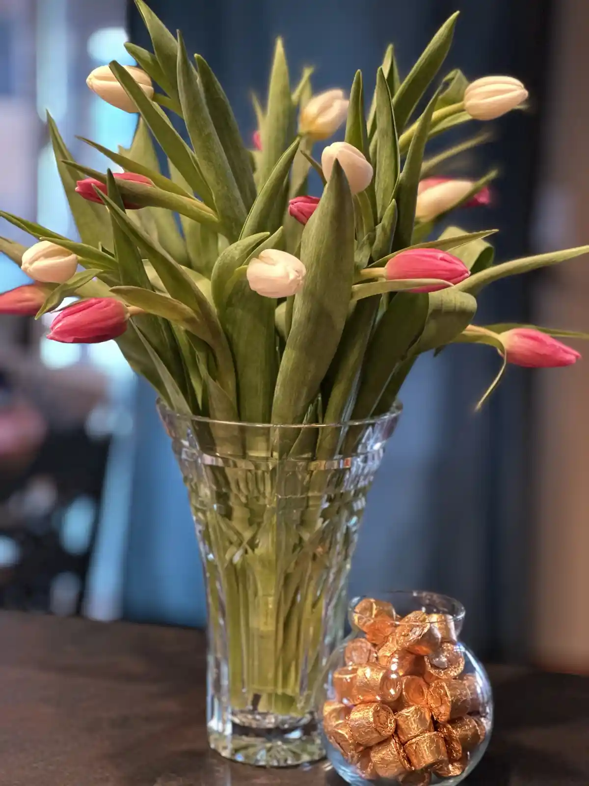 A clear glass vase filled with pink and white tulips sits on a dark surface next to a small glass bowl filled with…