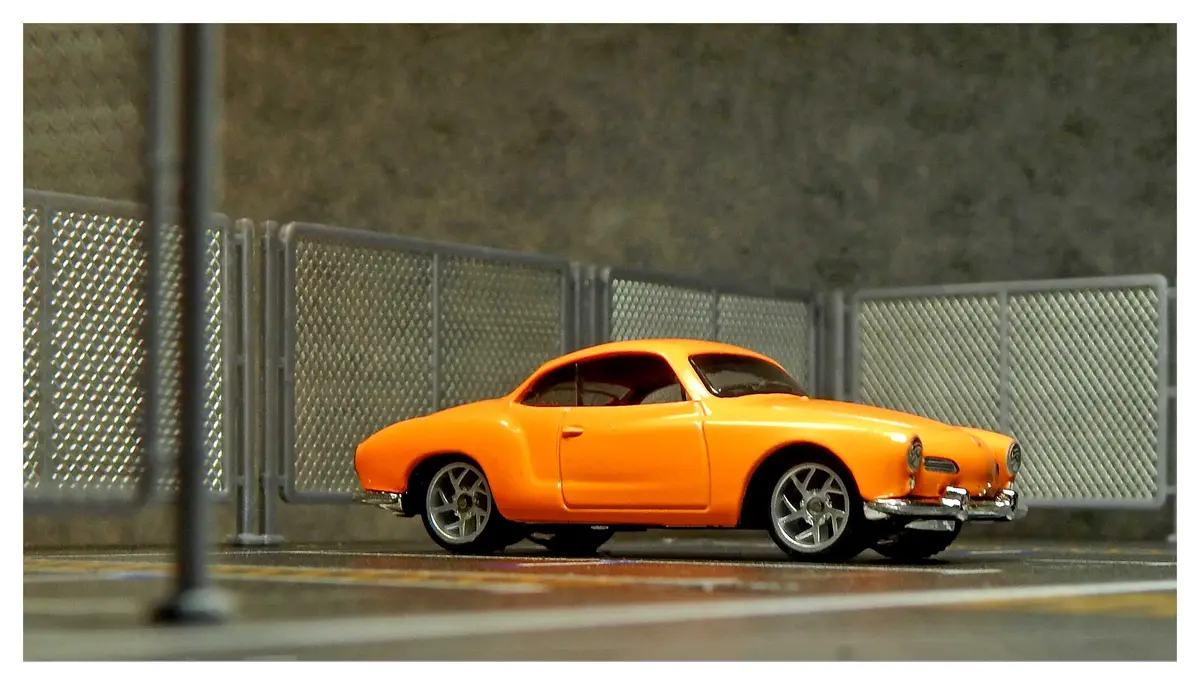 A bright orange toy model of a classic Volkswagen Karmann Ghia is parked on a miniature road next to a chain-link fence. The…
