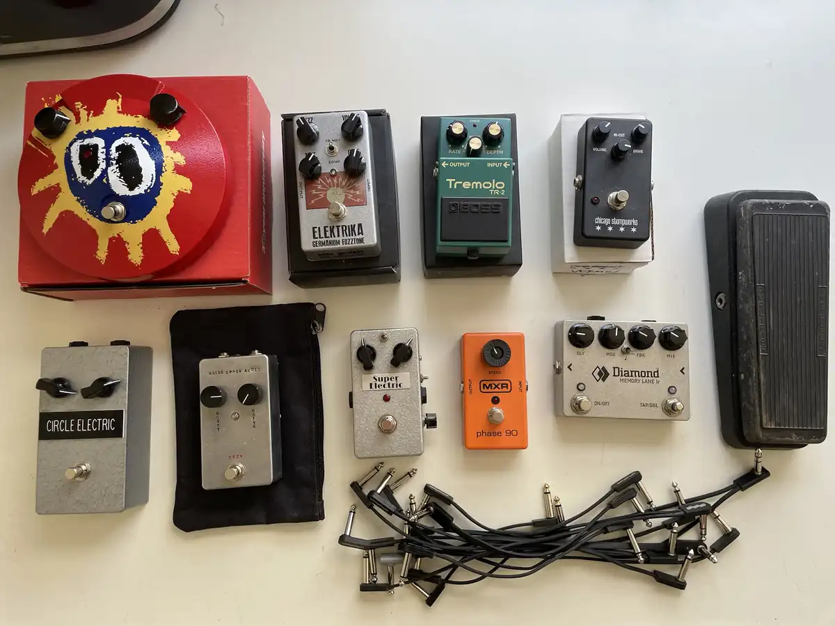 An overhead shot displays a collection of ten guitar effect pedals arranged on a white surface, alongside a pile of ten…