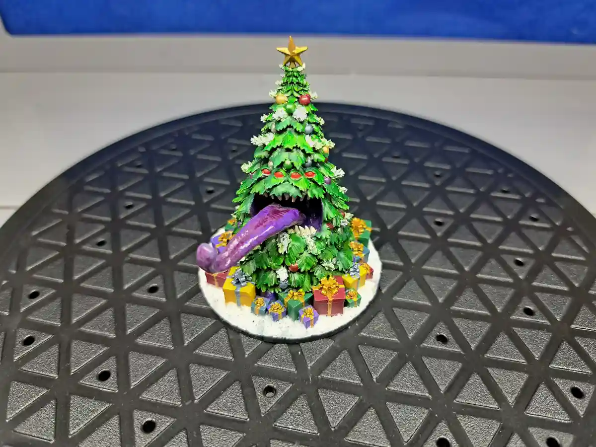 A miniature Christmas tree decorated with ornaments and presents sits on a black gridded surface, but its trunk is replaced…