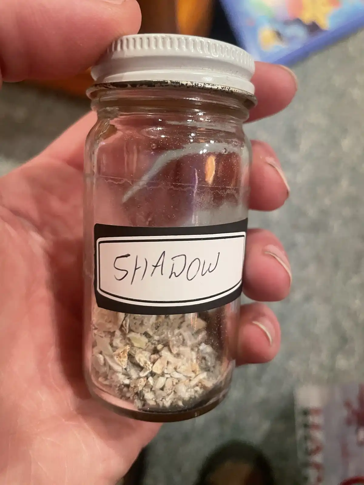 A hand holds a small glass jar with a white lid, containing a collection of light-colored, fragmented, granular material…