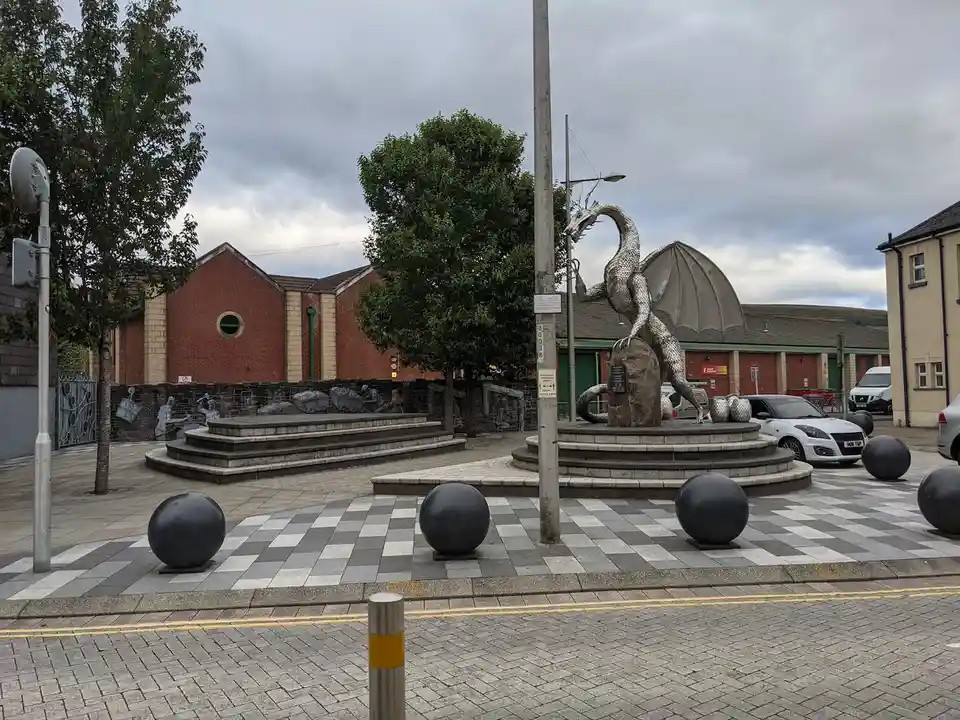 A large, metallic dragon statue is perched atop a stone pedestal in a public plaza, its head awkwardly stuck in the branches…
