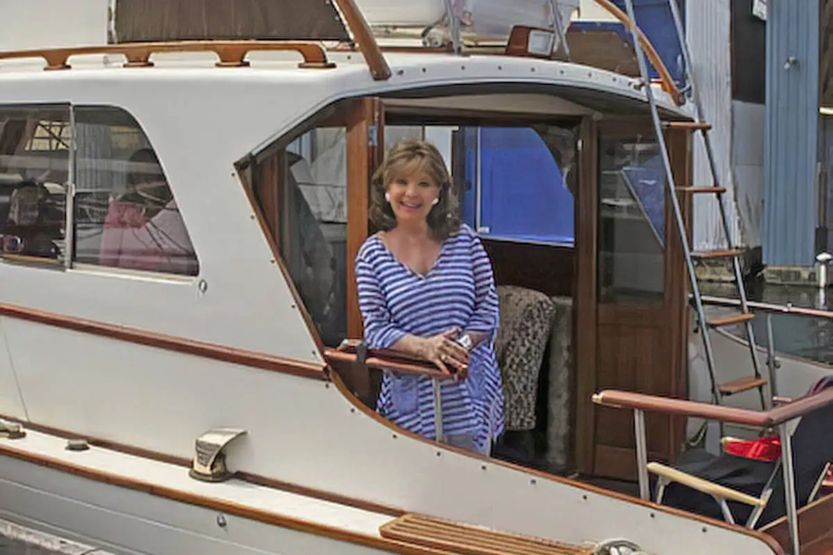 a woman in a blue and white striped shirt smiles while leaning on the railing of a white boat with wooden trim. The boat…
