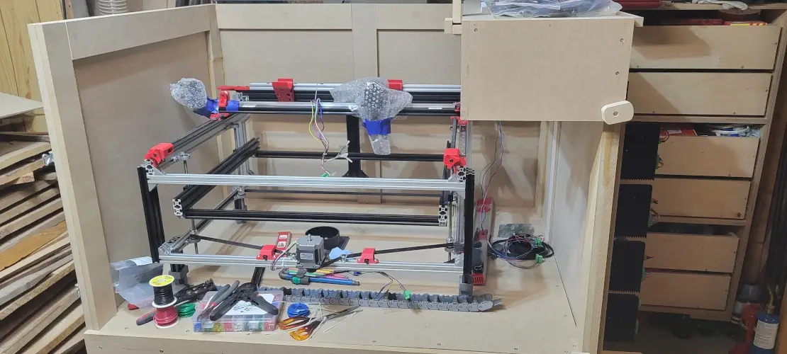 A partially assembled 3d printer sits inside a wooden enclosure, surrounded by tools, wires, and spools of filament. The…