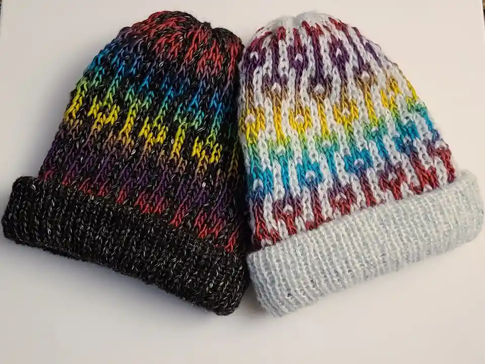 Two hand-knitted, colorful beanie hats with rainbow-striped patterns sit side by side on a white surface. The hat on the…
