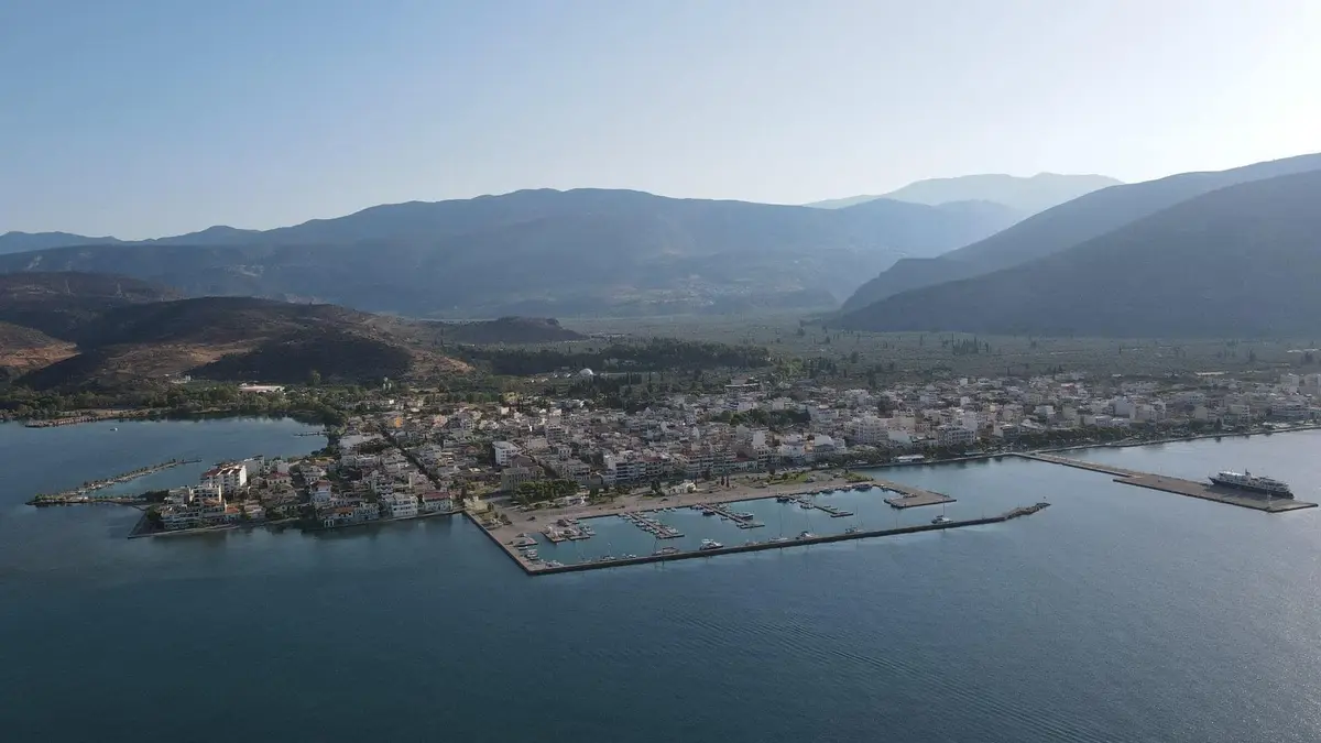 An aerial photo shows a coastal town nestled between mountains and water, with a harbor and boats visible. Overlaid text…