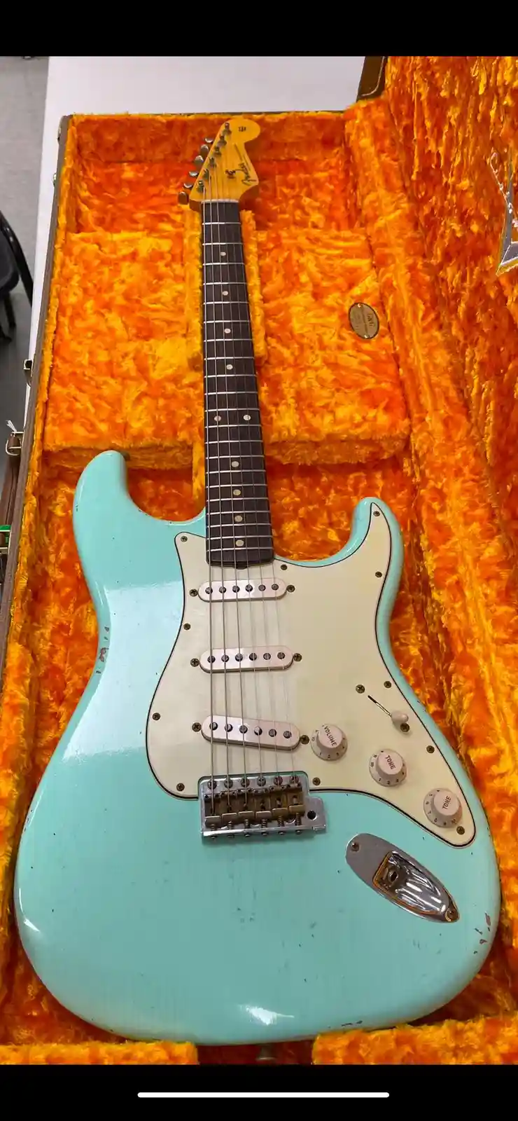 A light blue Fender Stratocaster electric guitar with a worn finish rests inside its open case, which is lined with bright…