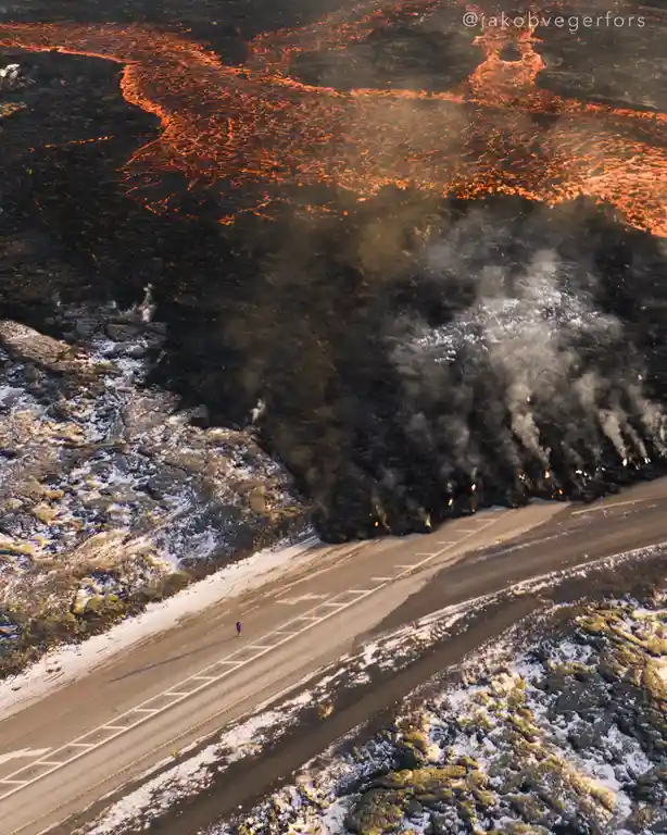 An aerial photo shows a lone person running on a snowy road next to a vast, glowing orange lava flow. The lava is actively…
