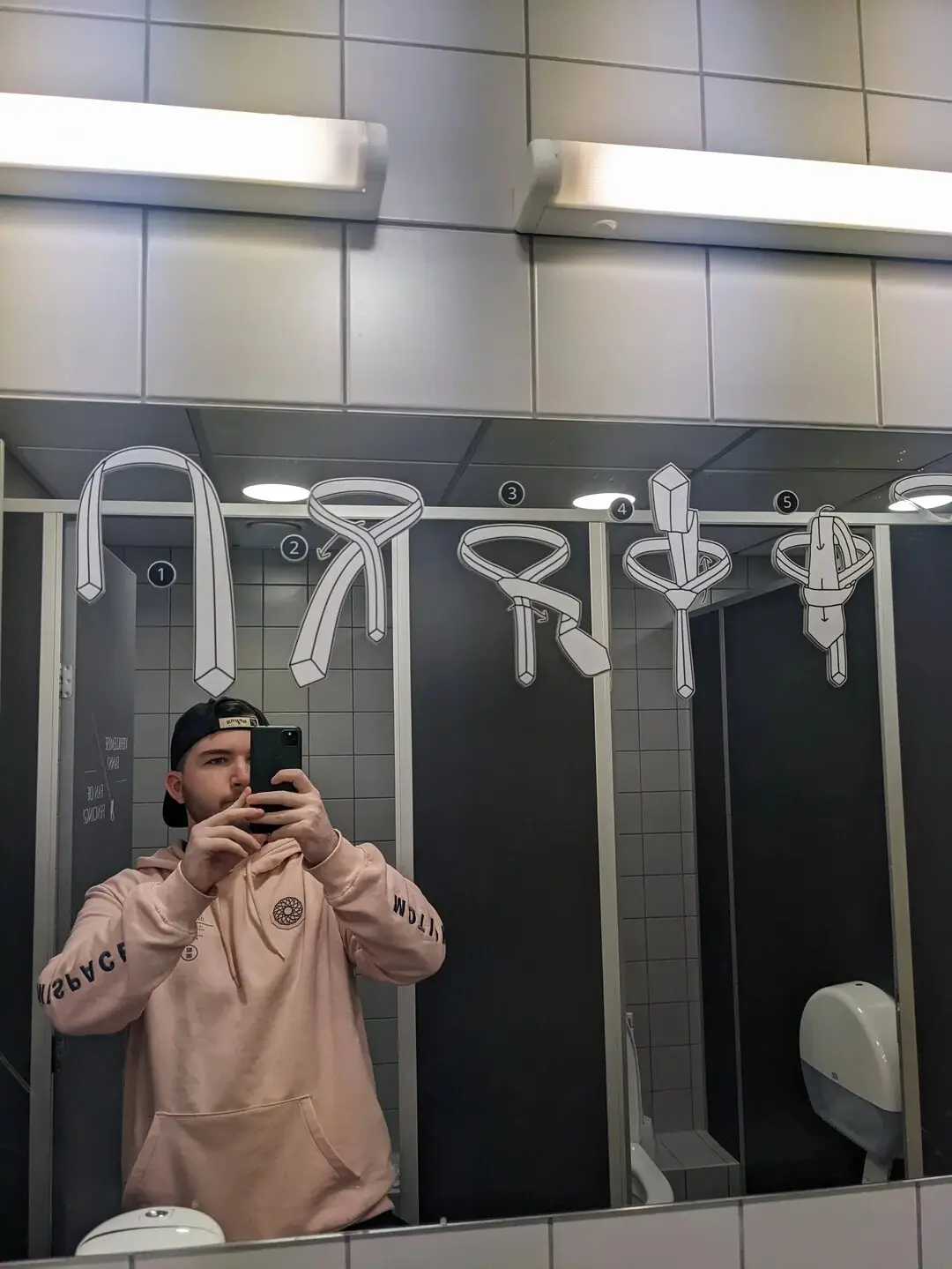 A man in a pink hoodie and black cap takes a mirror selfie in a public restroom, with five numbered illustrated tie knots…