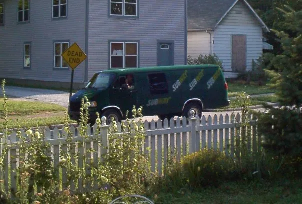 A green Suburban delivery van is parked on a residential street next to a white picket fence and houses. A yellow…