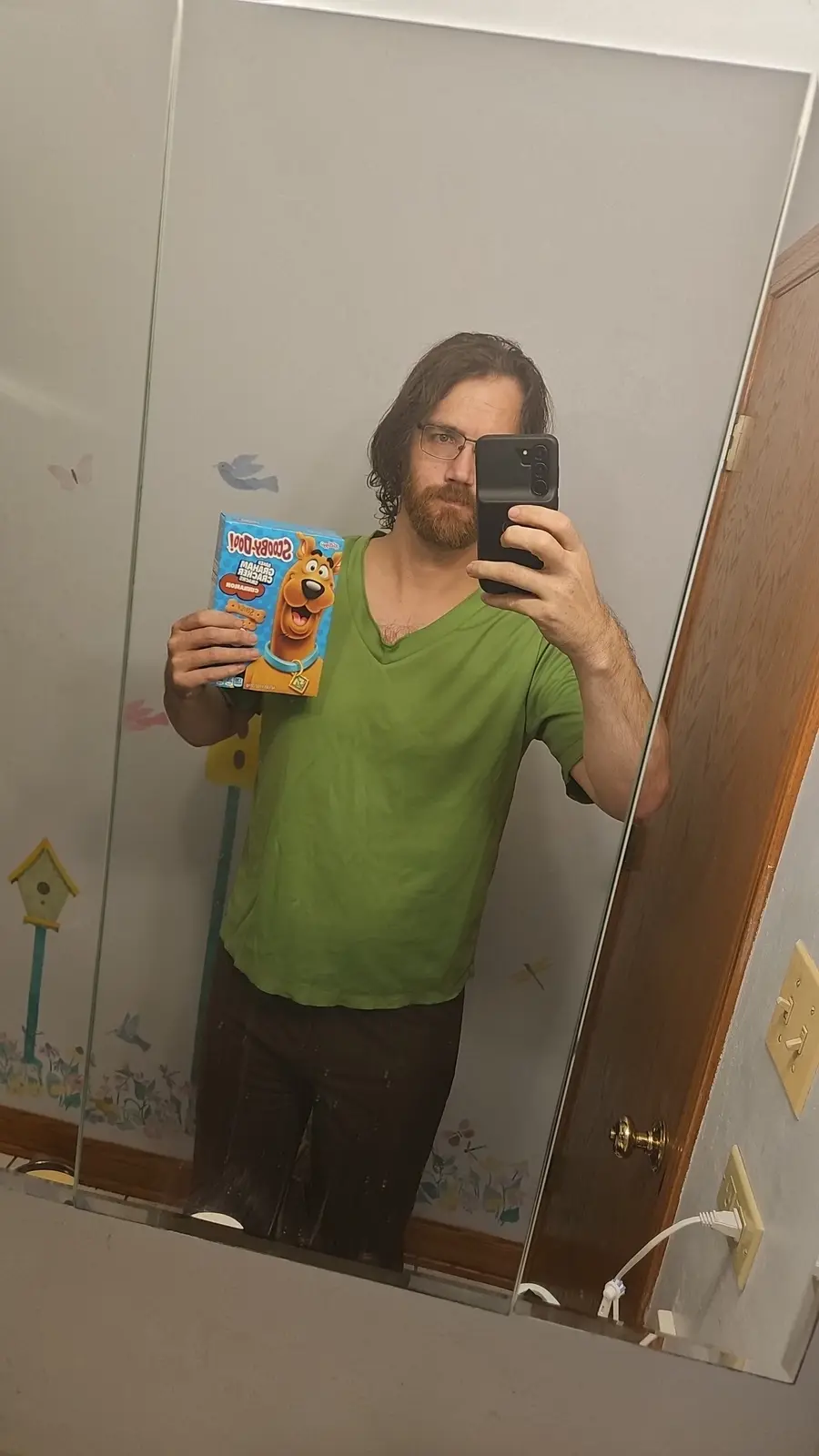 A man with glasses and a beard takes a mirror selfie in a bathroom, holding up a box of Scooby-Doo cereal while obscuring…