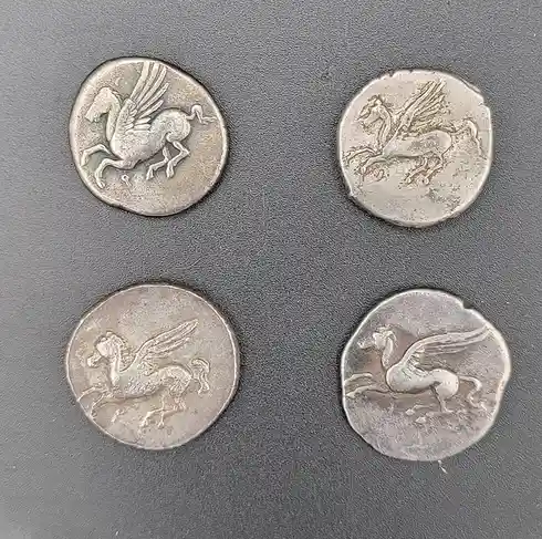 Four ancient silver coins are arranged in a square on a dark gray textured surface, each featuring a different stylized…