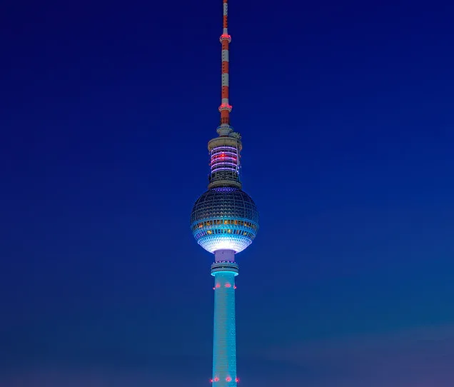 A nighttime photo of the Berlin TV Tower (Fernsehturm) illuminated with blue, red, and white lights against a dark blue sky…