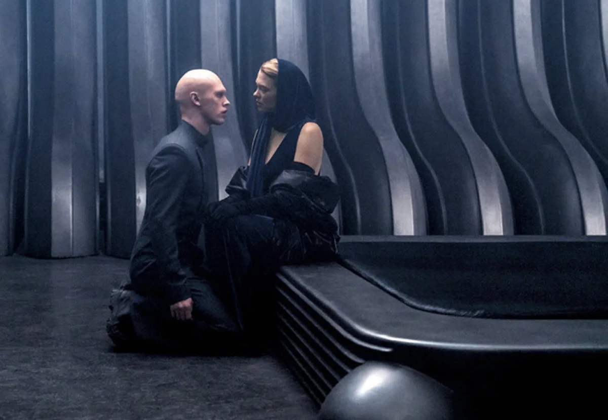 A bald man in black kneels on the floor facing a woman in black who sits on a sleek black bench against a wavy metallic…