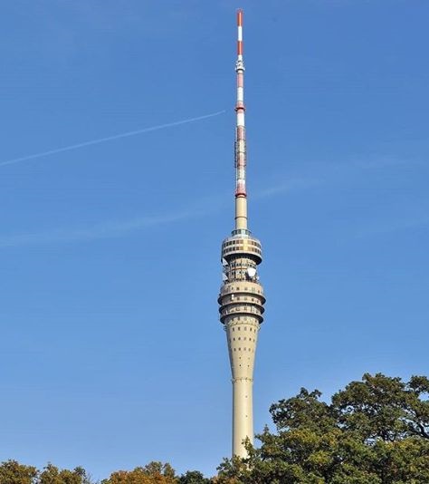 A tall, slender telecommunications tower with red and white stripes near the top stands against a clear blue sky, with tree…