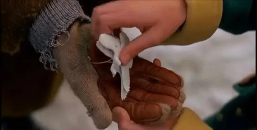 A close-up shot shows one person's gloved hand holding a small white paper bird while another person's bare hand gently…