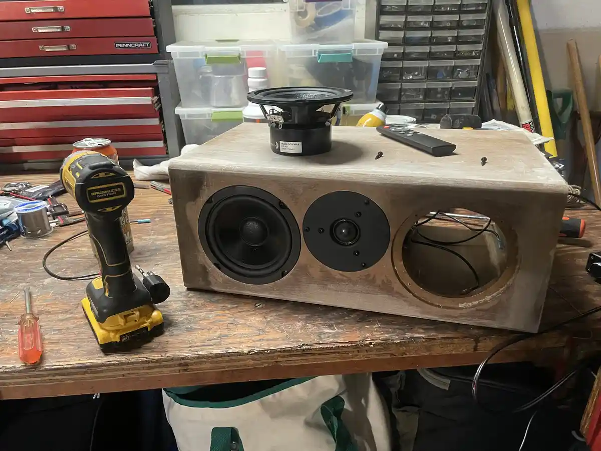 A homemade wooden speaker box sits on a cluttered workbench, featuring two installed drivers and an empty port. A third…