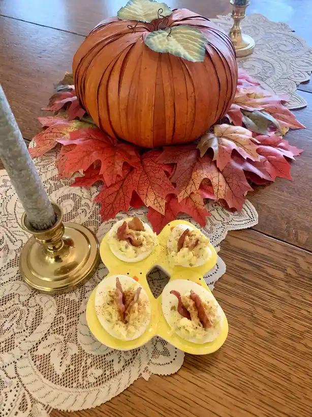 A yellow egg tray holding four deviled eggs with bacon bits and paprika sits on a lace doily next to a decorative pumpkin…
