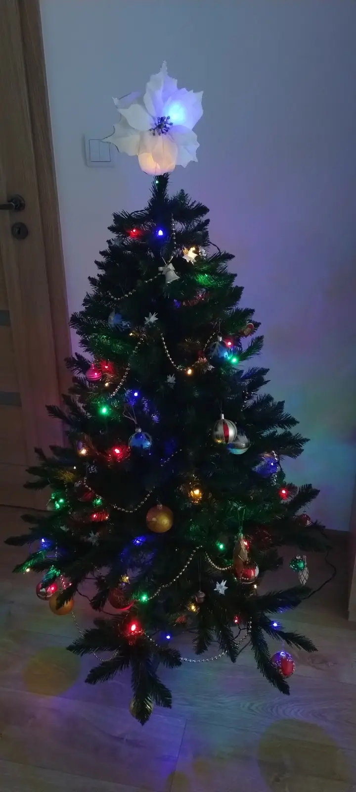 A decorated artificial Christmas tree with colorful lights and ornaments stands indoors, topped with a large white…
