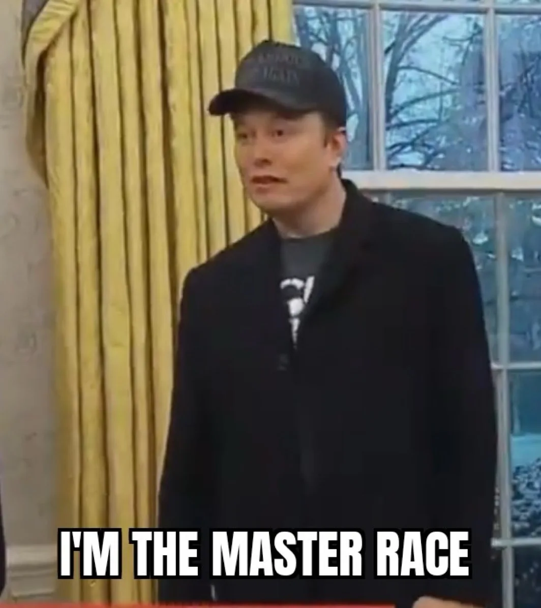 A man wearing a black cap and black coat stands in front of yellow curtains and a window with bare trees visible outside…