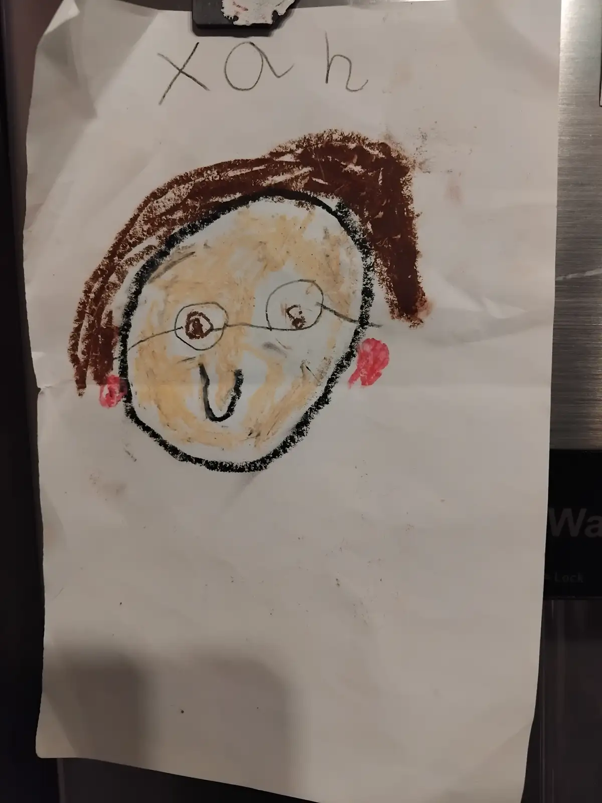 A child's crayon drawing on paper shows a face with brown hair, glasses, red cheeks, and a smiling mouth, with the name…