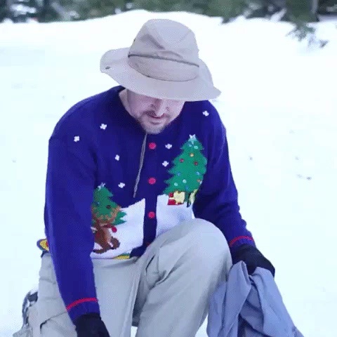 A man in a beige hat and light pants kneels in snow, visibly uncomfortable in a bright blue Christmas-themed sweater with…