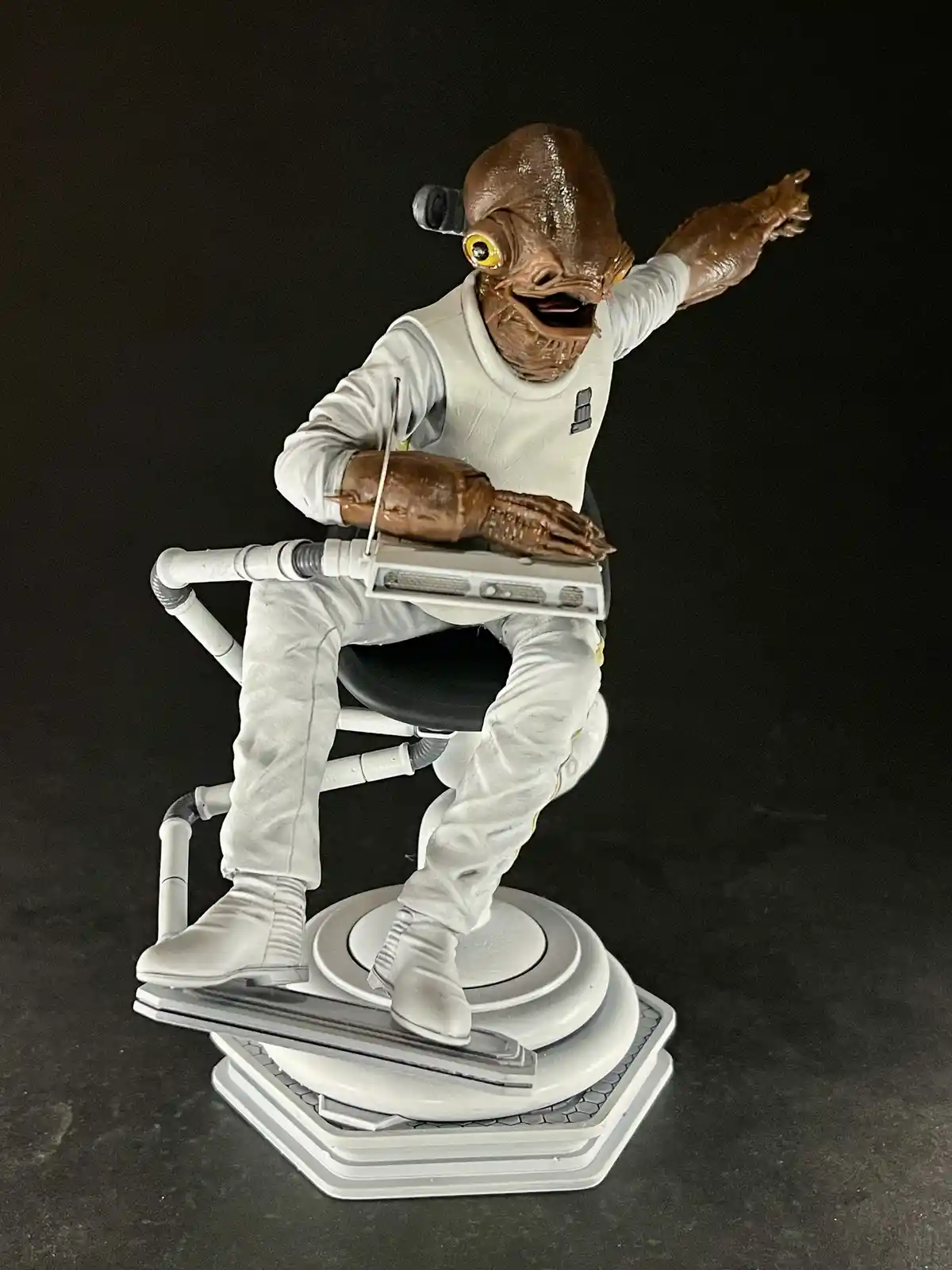 A detailed figurine of a brown-skinned, amphibious alien with large yellow eyes, wearing a white uniform and seated on a…