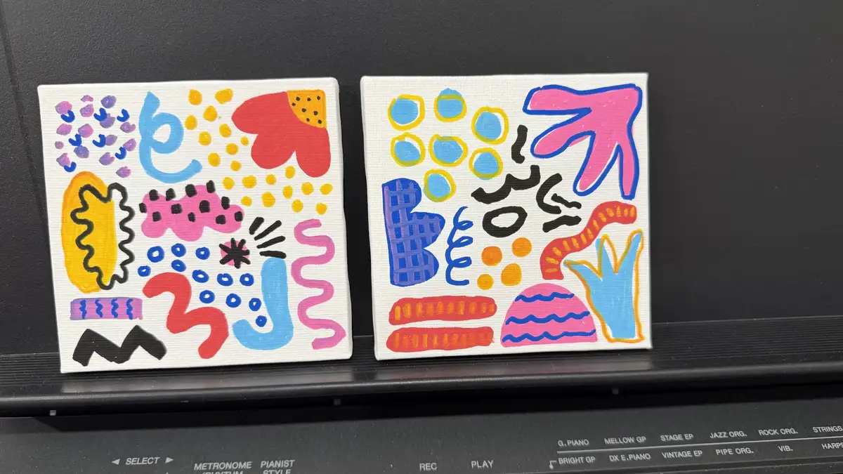 Two small, colorful abstract paintings with playful shapes and patterns sit side-by-side on a black electronic keyboard. The…