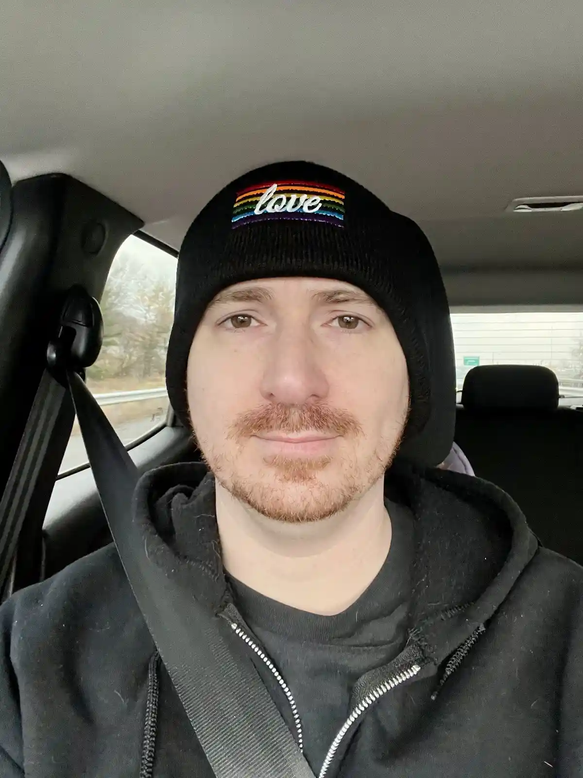 A man with a light beard and brown eyes sits in a car wearing a black beanie with a rainbow-colored 'love' patch and a black…