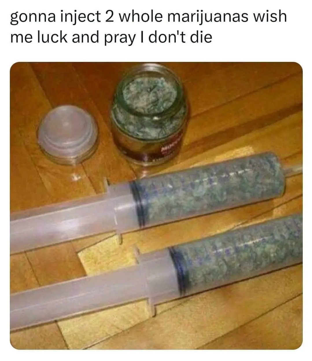 Two large syringes filled with green marijuana buds lie on a wooden floor next to an open glass jar containing more buds and…